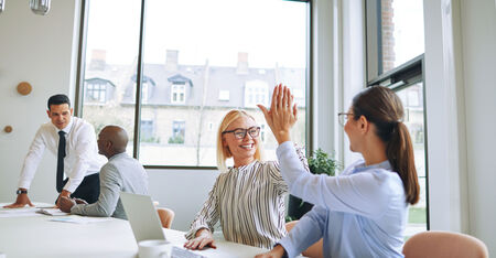 Zwei freundliche Projektmanagerinnen die sich ein Highfive geben, im Hintergrund sind Mitarbeiter 